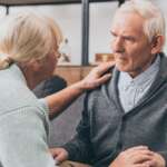 A petite senior woman holding and stroking her husband shoulder, trying to console him, while he is in different stages of dementia