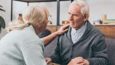 A petite senior woman holding and stroking her husband shoulder, trying to console him, while he is in different stages of dementia