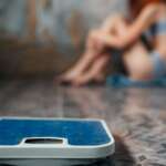A close-up image of weighing machine and a young female in a blurry background feeling fear of weight gain representing anorexia nervosa