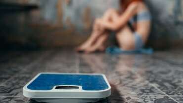 A close-up image of weighing machine and a young female in a blurry background feeling fear of weight gain representing anorexia nervosa