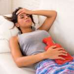 A sad woman is laying on couch with a hot water bottle on her abdomen due to irregular periods
