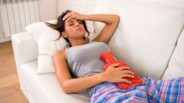 A sad woman is laying on couch with a hot water bottle on her abdomen due to irregular periods