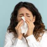 sick woman sneezes, isolated over purple wall, holding napkin showing the concept of influenza and how it triggers pneumonia