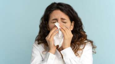 sick woman sneezes, isolated over purple wall, holding napkin showing the concept of influenza and how it triggers pneumonia