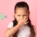 A little girl in a t-shirt is keeping hand on mouth and looking disgusted due to smelly fart around