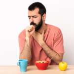 Caucasian man having breakfast at a table coughing a lot, showing the signs of food allergies and management tips.