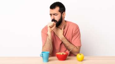 Caucasian man having breakfast at a table coughing a lot, showing the signs of food allergies and management tips.