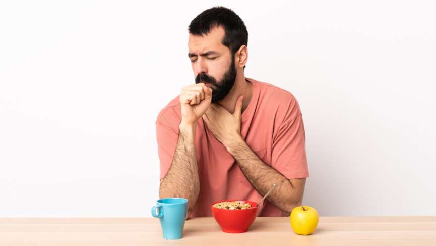 Caucasian man having breakfast at a table coughing a lot, showing the signs of food allergies and management tips.