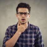a young man sits, holding his throat, feeling a sore, burning throat, representing vitamin b12 deficiency concept