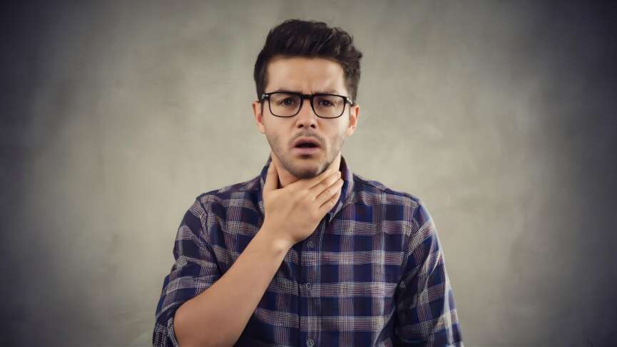 a young man sits, holding his throat, feeling a sore, burning throat, representing vitamin b12 deficiency concept