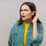 A curious young pretty Caucasian woman is keeping her hand behind her ear, doing the "I can't hear you" gesture. causes of hear loss concept