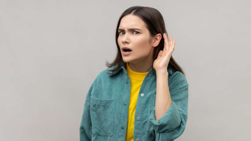 A curious young pretty Caucasian woman is keeping her hand behind her ear, doing the "I can't hear you" gesture. causes of hear loss concept