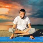 fitness, sport, yoga, people and healthy lifestyle concepts—a man making scale pose lotus variation on mat outdoors on sea pier over sunset background for improving fertility