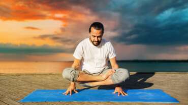 fitness, sport, yoga, people and healthy lifestyle concepts—a man making scale pose lotus variation on mat outdoors on sea pier over sunset background for improving fertility