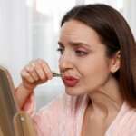 A pretty young lady with hair removal plucking her upper lip near a mirror. Hirsutism and excess body hair concept