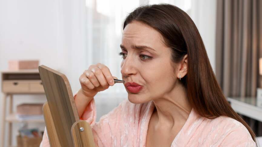 A pretty young lady with hair removal plucking her upper lip near a mirror. Hirsutism and excess body hair concept