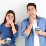 A young couple posing in a white wall having contagious yawn, showing a tired gesture, and covering their mouth with hands