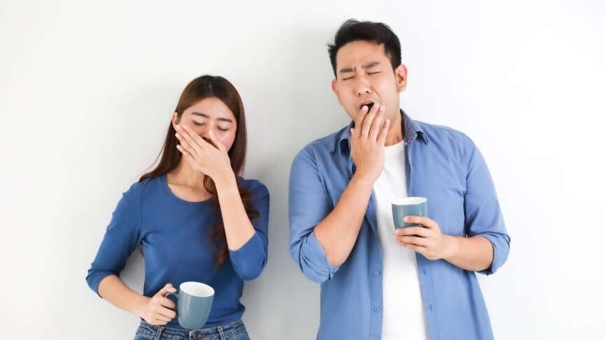 A young couple posing in a white wall having contagious yawn, showing a tired gesture, and covering their mouth with hands