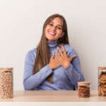A young Caucasian woman sitting at a table with a food pot for breast cancer prevention, isolated on a white background, has a friendly expression, pressing her palm to chest.