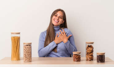 A young Caucasian woman sitting at a table with a food pot for breast cancer prevention, isolated on a white background, has a friendly expression, pressing her palm to chest.