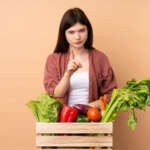 Young farmer girl with freshly picked vegetables in a box frustrated and pointing to the front, representing disadvantages of a plant-based diet