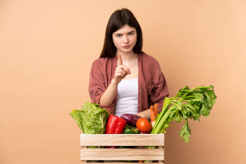 Young farmer girl with freshly picked vegetables in a box frustrated and pointing to the front, representing disadvantages of a plant-based diet